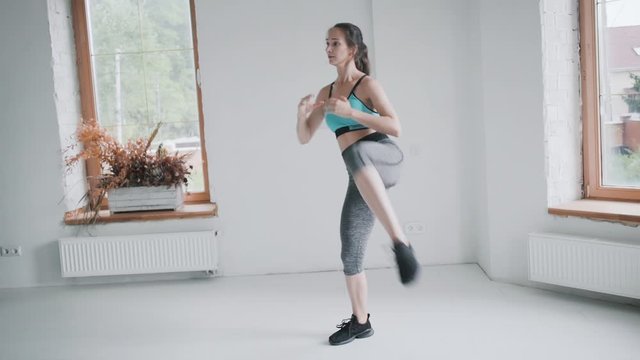 Young fit brunette girl with pigtail in blue top, gray leggings , black sneakers does exercises in bright room. Doing sports and fitness training at home. Healthy lifestyle. girl makes lunges, punches