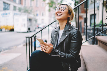 Cheerful asian hipster girl looking up laughing at joke read in social network while sitting on stairs in downtown, happy Chinese female using application on smartphone for chatting, receive message