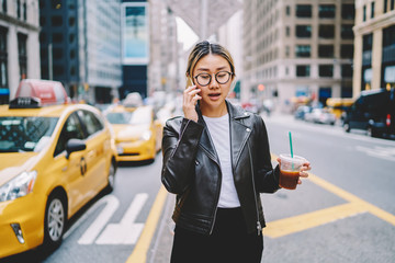 Young asian woman in eyewear walking with ice coffee cup calling taxi contact service operator on...