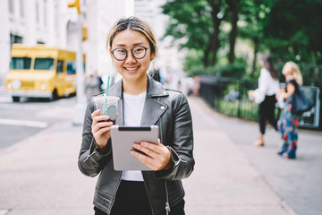 Half length portrait of charming emotional asian female blogger enjoying recreation with digital tablet and tasty drink.Cute cheerful woman looking at camera and laughing while walking at street