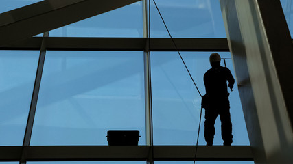 Worker man is washing windows with scraper on a highrise modern building hanging the rope, close-up