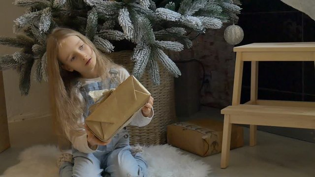 Cute blond girl has recieved a gift box wrapped in craft paper sitting near christmas tree, happy childhood and xmas celebration concepts