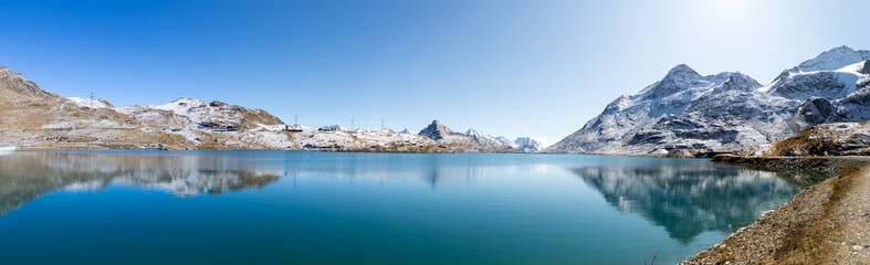Obraz na płótnie Canvas Panoramic view of Lago Bianco at Bernina pass. Lake reflects mountain range covered in snow and the blue sky. Picture taken after midday in late Septemeber