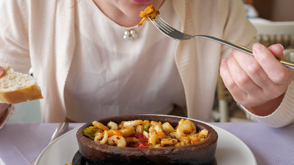 Good served food in restaurant. Unrecognizable woman is eating frying hot shrimps with vegetables served in clay bowl, closeup dish. Takes shrimp out of the boiling oil with a fork.