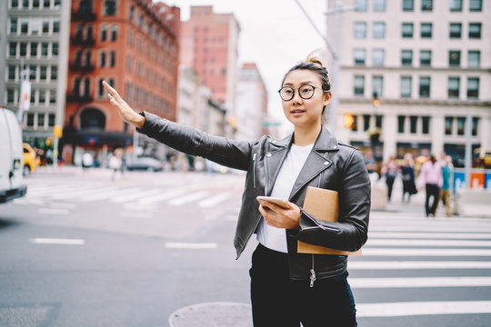 Young attractive asian woman trying to catch taxi while using online GPS-tracker application on smartphone connected to 4G.Cute hipster girl in eyewear gesturing for stop public transport at street