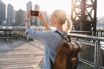 Back view of millennial woman shooting video of New York landscape using smartphone for clicking...