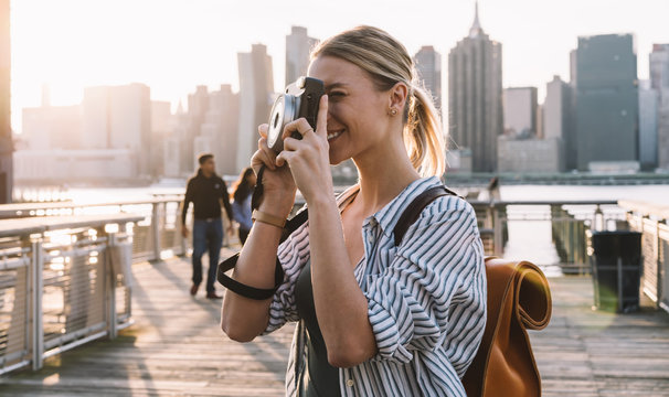 Young female tourist with stylish rucksack standing at urban setting with Manhattan district on background and clicking pictures with camera for get few instant photos at moment