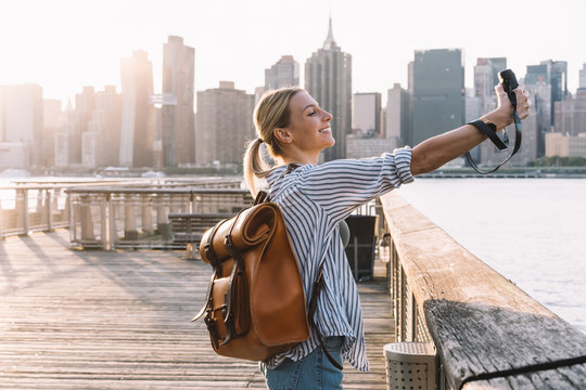 Side view of cheerful woman traveller with touristic backpack enjoying American getaway posing for clicking selfie picture with retro instant camera, concept of happiness and USA vacations