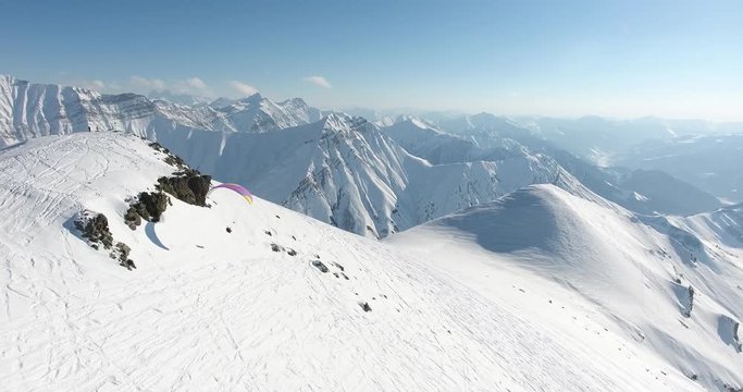 Paragliding sport in winter season. Parachute sky-diver flying over high mountains with fresh snow on sunny winter day. Top aerial  view. Caucasus Mountains, Georgia, ski resort Gudauri.