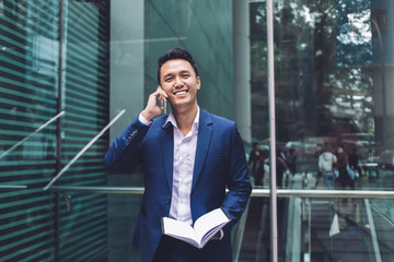 Delighted young modern Asian businessman talking over smartphone on street