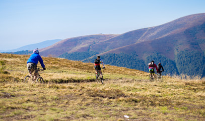 Obraz premium Group of unrecognizable tourists friends ride bicycles along top of mountains road in scenic green valley. Mountain bikers cruise through picturesque nature on sunny day.