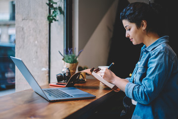 Caucasian female blogger writing content information for own website while sitting with laptop device at cafeteria table, attractive woman preparing to course work indoors holding textbook