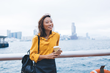 Obraz premium Half length portrait of cheerful female student with takeaway cup smiling at camera while waiting friend at Hong Kong urban setting for exploring Chinese metropolis, positive blogger rejoicing outdoor