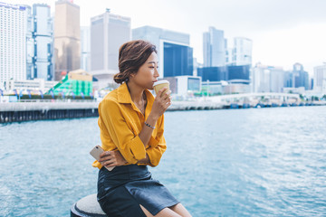 Pensive female tourist drinking tasty caffeine beverage from takeaway cup during rest time on city area with Hong Kong on background, contemplative Japanese woman recreating during vacations