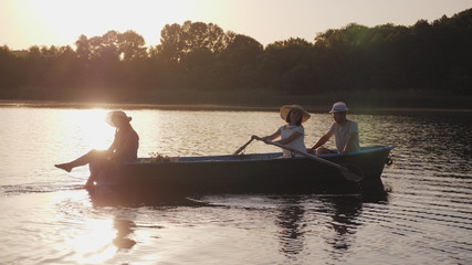 Young family floating in a boat with oars at sunset. Slow motion. Girl in a hat smiling, rowing paddles, teen daughter sitting at the stern of wooden boats and wets feet in water. Side view - Powered by Adobe