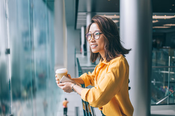 Young Asian woman with coffee and cellphone leaning on railing smiling and daydreaming - Powered by Adobe