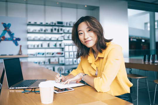 Portrait of positive hipster student sitting at desktop with knowledge textbook and laptop for e learning and smiling at camera, successful female freelancer working remotely with distance plan