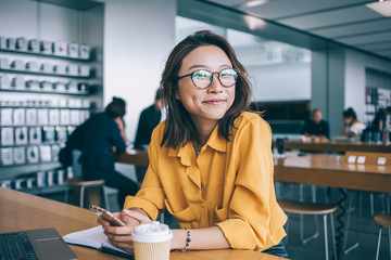 Fototapeta Portrait of cheerful hipster girl in optical spectacles for provide eyes protection looking at camera and smiling during blogging time in university classroom, concept of browsing and networking obraz