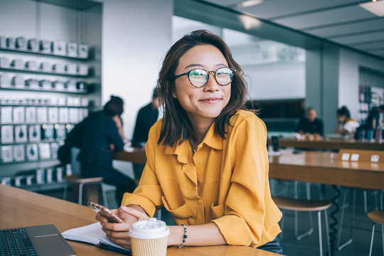 Portrait of cheerful hipster girl in optical spectacles for provide eyes protection looking at camera and smiling during blogging time in university classroom, concept of browsing and networking