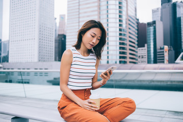 Chinese millennial blogger networking website during free time at city street connected to 4g wireless on cellphone gadget, attractive Japanese female generation typing content text for sharing to web