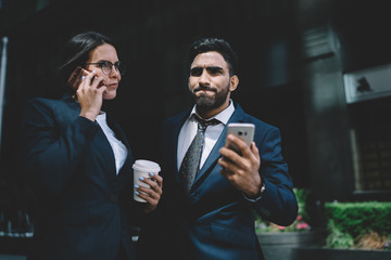 Serious woman in formal wear making international call for discussing details of contract with business partner while spanish male colleague standing near and feeling puzzled on notification of mobile