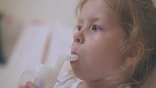 pretty blue eyed little girl breathes with modern inhaler lying in comfortable bed in light room extreme close view