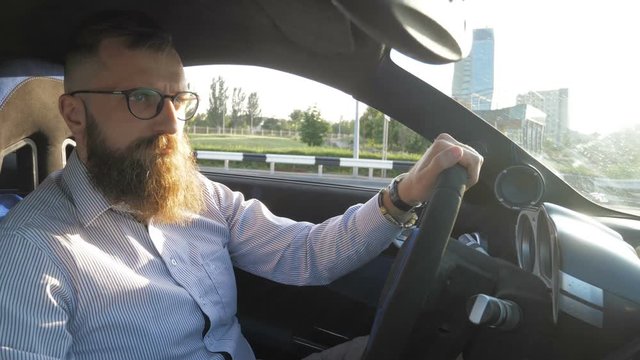 Serious young bearded man in eyeglasses sitting behind the wheel and driving sport car