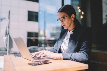 Concentrated businesswoman chatting online with partners using internet connection on laptop computer during time in street cafe, elegant caucasian banker working with documents on netbook