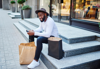 Portrait of afro american guy waiting for feedback from web store service sitting on stairs waiting for purchase via smartphone, positive dark skinned man in stylish hat resting after shopping time