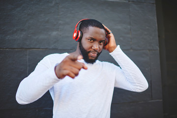 Portrait of young man pointing on you and looking at camera and listening music via modern stereo headphones, handsome hipster guy in trendy casual apparel enjoying radio via new equipment outdoors