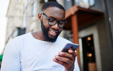 Positive millennial man chatting online on smartphone spending time outdoors in urban setting,...