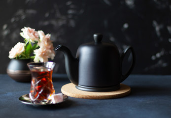 Black teapot, traditional eastern armudu glass of tea and flowers on dark background. Copy space