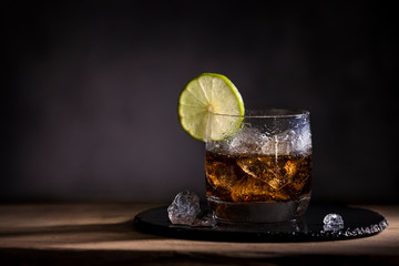 A glass of ice cold refreshing coke with a slice of lime as a decoration on a round slate plate on a wooden table.