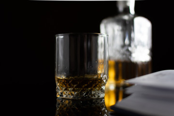 A glass of whiskey close-up and a business diary on a black glass table on a black background. Blurred decanter of whiskey in the background