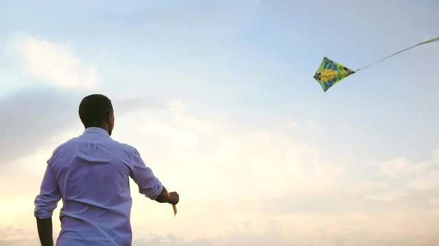 young man in white shirt flies colorful kite in sky with light clouds at sunset backside low angle shot slow motion