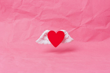 Fototapeta premium Happy valentines day banner. A red wooden heart with white voluminous wings soars over a pink crumpled paper background. Minimalism. Place for text