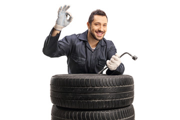 Cheerful auto mechanic gesturing great with hand and holding wrench behind car tires