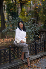 Autumn fashion outdoor. The girl in fashionable stylish white shirt , pants and sunglasses, autumnal lifestyle on the background of blurry yellow-green trees in the park. Vertical