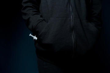 a man holds his hands in the pockets of a sweatshirt and a syringe with a drug peeks out of one pocket