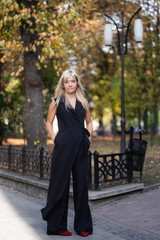 Autumn fashion outdoor. The blonde girl in fashionable stylish black overalls, autumnal lifestyle on the background of blurry yellow-green trees in the park. Vertical