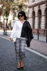 Fototapeta premium Autumn fashion outdoor. The girl in fashionable stylish white shirt , pants, jacket and sunglasses, autumnal lifestyle on the background of blurry yellow-green trees in the park. Vertical