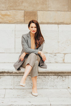 Outdoor Portrait Of Beautiful Woman Resting In Park, Sitting On Bench, Wearing Checked Trousers And Coat