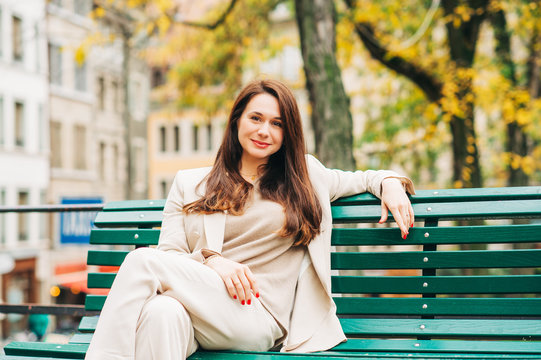 Outdoor portrait of beautiful woman resting in park, sitting on bench, wearing beige suit