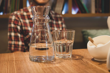 cropped view of man in checkered shirt sitting at wooden table with sugar bowl, glass and jud with water in cafe