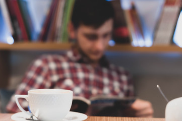 soft focus of young man in checkered shirt sitting and reading magazine near table with white cup in front of shelves with books in cafe