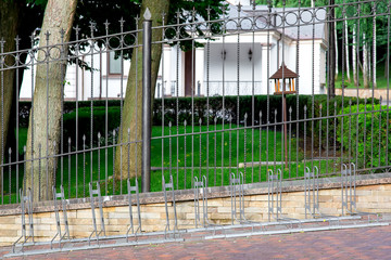 empty bike parking on the sidewalk made of tiles near a black metal fence with peaks in a park with trees in the background plants and a light building..