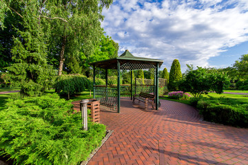 Fototapeta premium an iron gazebo with a bench and paving stone tiles in the garden with a lawn and evergreen thuja on a sunny day with a cloudy sky, nobody.