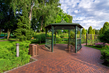 an iron gazebo with a bench and paving stone tiles in the garden with a lawn and evergreen thuja near the canopy of the urn and in the bushes an iron lantern on a sunny day with a cloudy sky, nobody.