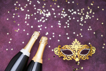 Two champagne bottles, golden carnival mask and confetti stars on purple background. Flat lay of...