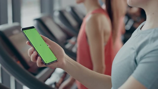 
Athletic Woman Uses Green Mock-up Screen Smartphone while Running on the Treadmill in a Gym. Sports Female Posts on Social Media, Takes Pictures, Watches Videos while Exercising in the Fitness Club